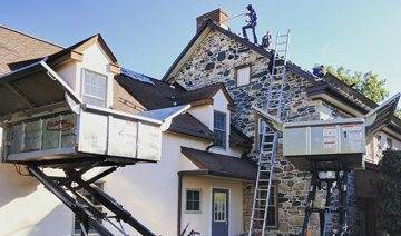 caution tape, machinery, and ladder in front of house with roofers on the roof